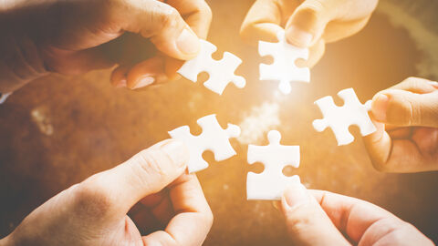 Symbolbild Partner Fünf Hände halten fünf Puzzleteile kreisförmig aneinander. Die Puzzleteile sind hell ausgeleuchtet.