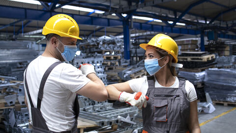 Kollegin und Kollege begrüßen sich coronakonform Ein Arbeiter und eine Arbeiterin in einer Industriehalle mit Masken, gelben Sicherheitshelmen und Arbeitslatzhosen begrüßen sich coronakonform per Ellbogencheck.