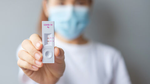 Symbolbild Corona-Schnelltest Scharf im Vordergrund ist ein positiver Corona-Selbsttest zu sehen, den eine Frau in die Kamera hält. Sie ist nur unscharf im Hintergrund zu sehen und trägt eine OP-Maske und ein weißes T-Shirt.
