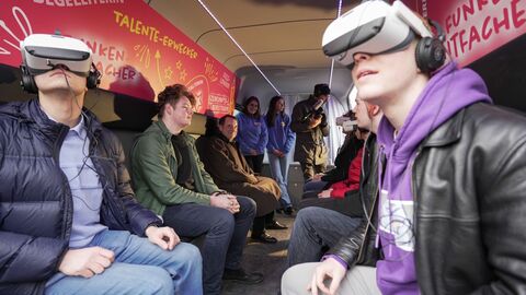 Mehrere Jugendliche mit VR-Brillen sitzen sich gegenüber auf Bänken in einem Bus, Kultusminister Lorz im Hintergrund beobachtet die Jugendlichen