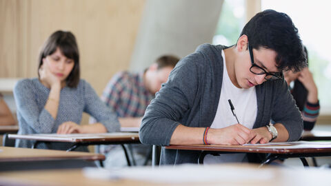 Ein junger Mann sitzt an einem einzelnen Tischchen und schreibt in seine Unterlagen, im Hintergrund weitere Personen über ihre Unterlagen gebeugt.