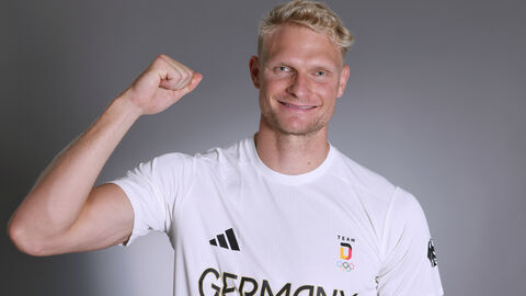 Oliver Zeidler Ein blonder Mann mit weißem T-Shirt streckt den arm hoch und hat einen begeisterten Gesichstausdruck