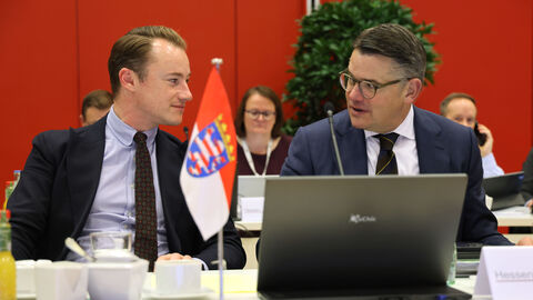 Chef der Staatskanzlei, Benedikt Kuhn und Ministerpräsident, Boris Rhein. Zwei Männer sitzen in Anzügen nebeneinander und schauen sich an. Vor ihnen steht auf dem Tisch ein Laptop.