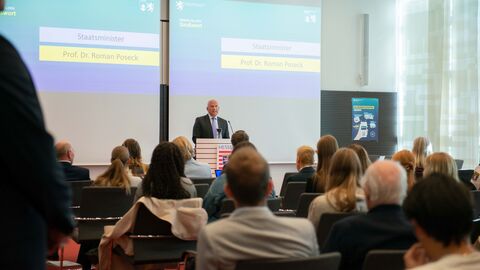 Innenminister Roman Poseck begrüßt die Schülerinnen und Schüler der Gewinnerteams im Innenministerium in Wiesbaden. Roman Poseck steht an einem Pult und hält vor einem Publikum eine Rede.