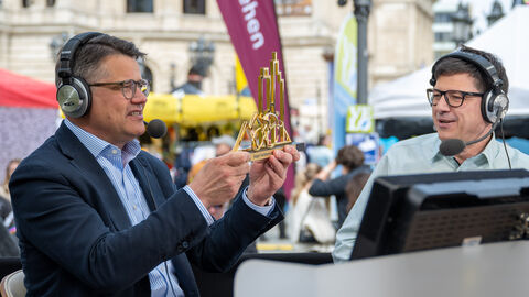 Ministerpräsident Rhein mit Kofhörern im Gespräch mit einem anderen Mann mit Kopfhörern