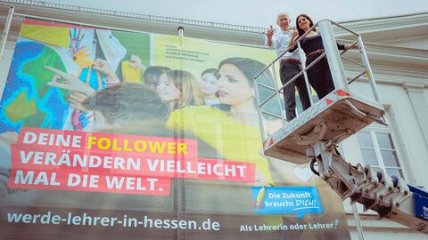 Von rechts auf einem Kran steht Kultusminister Schwarz gemeinsam mit einer Dame, hintendran an der Wand ist ein Plakat zu sehen mit der Aufschrift: "Deine Follower verändern vielleicht mal die Welt." der Initiative „Die Zukunft braucht Dich! Als Lehrerin oder Lehrer“.