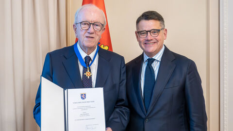 Ministerpräsident Boris Rhein überreicht Dr. Elmar Reiss aus Frankfurt am Main den Hessischen Verdienstorden. Zwei Männer stehen nebeneinander. Der linke hält eine Urkunde hoch.