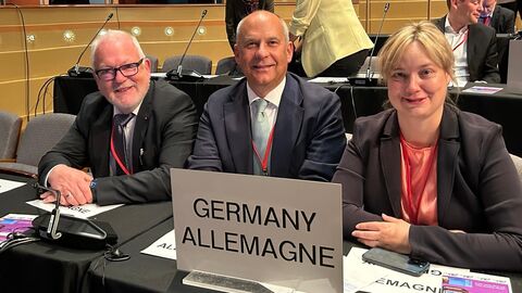 Zwei Männer und eine Frau schauen in Kamera. Auf dem Schreibtisch vor ihnen ist ein Schild mit der Aufschrift "GERMANY ALLEMAGNE"