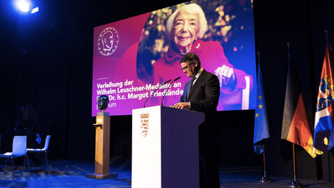 Person steht an Rednerpult. Auf dem Bildschirm im Hintergrund sieht man ein Foto einer älteren Frau.